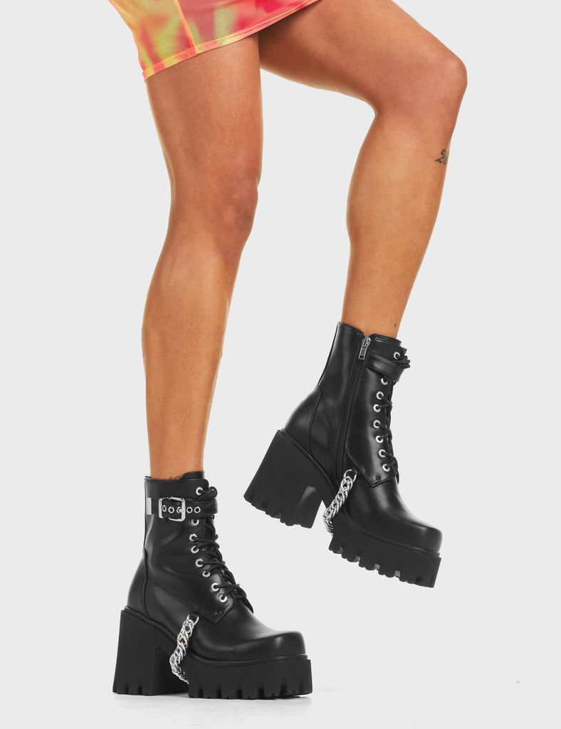 Play Hard Chunky Platform Ankle Boots in Black. Features lace-up detailing, an adjustable strap with a silver square buckle, and hanging silver chain around the sole.